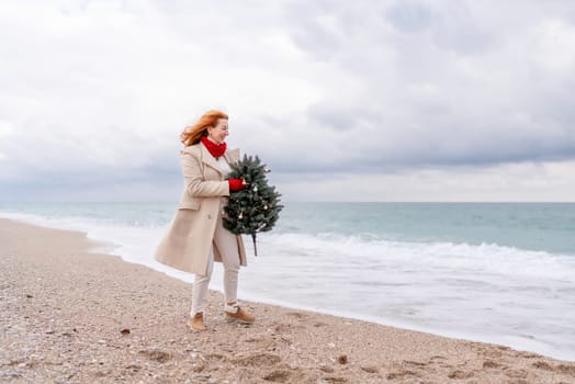 Redhead woman Christmas tree sea. Christmas portrait of a happy redhead woman walking along the beach and holding a Christmas tree in her hands. Dressed in a light coat, white suit and red mittens