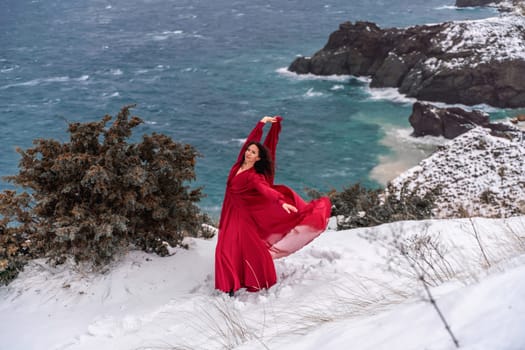 Woman red dress snow sea. Happy woman in a red dress in the snowy mountains by the emerald sea. The wind blows her clothes, posing against sea and snow background