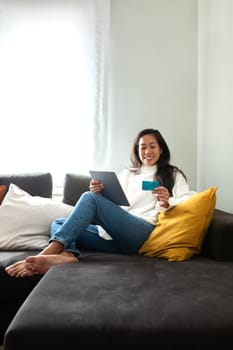 Young Asian woman relaxing on the couch shopping online using digital tablet and credit card at home. Copy space. Vertical image. E-commerce concept.