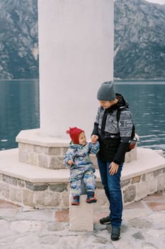 Mom stands near a little girl sitting on the steps of a lighthouse on a pier by the sea. High quality photo