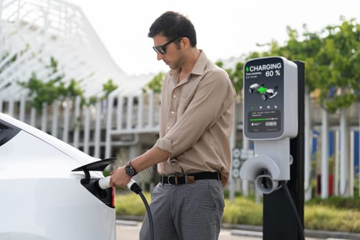 Young man put EV charger to recharge electric car's battery from charging station in city commercial parking lot. Rechargeable EV car for sustainable environmental friendly urban travel. Expedient