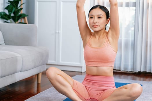 Asian woman in sportswear doing yoga exercise on fitness mat as her home workout training routine. Healthy body care and calm meditation in yoga lifestyle with comfortable and relaxation. Vigorous