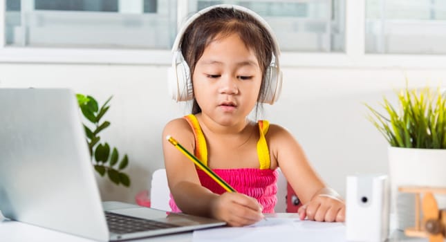 Little kid preschool wear headphone sit at desk use laptop computer and communicates on Internet online video call with teacher, Asian child girl study video conference distant education at home