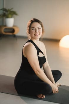 A girl in black sportswear is sitting on a yoga mat and smiling.
