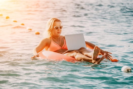 Woman works on laptop in sea. Freelancer, young blond woman in sunglases floating on an inflatable big pink donut with a laptop in the sea at sunset. Freelance, travel and holidays concept