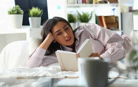Beautiful woman lies in bed and reads book. Home leisure and reading novels