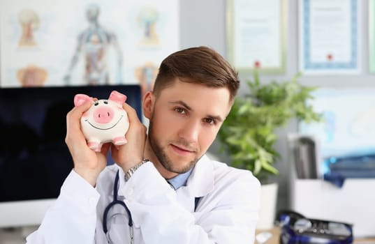 Portrait of male doctor holding a piggy bank on voluntary donations of money for a hospital or clinic. General practitioner collects charitable donation for patient care healthcare concept