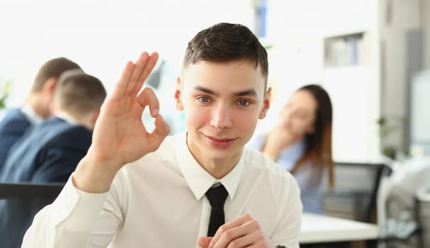 Young successful businessman shows good gesture on background of colleagues in office. Financial successful career growth in business