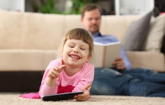 Little smiling baby girl with smartphone lies on floor in background dad reads a book. Children interests in phone and reading books