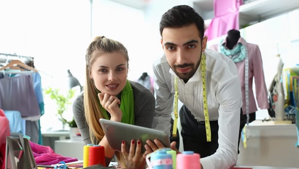 Man and woman designers are discussing new collection of clothes on digital tablet in workshop. Workers of online fashion store in modern warehouse.