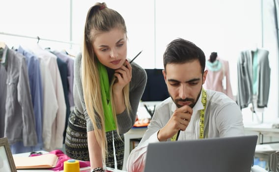 Two fashion designers work as a team at table and look thoughtfully at computer monitor. Teamwork in creative fashion atelier workshop