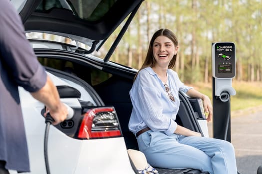 Lovely young couple recharging battery for electric car while relaxing and sitting on the trunk during road trip travel EV car in autumnal forest. Eco friendly travel on vacation during autumn. Exalt