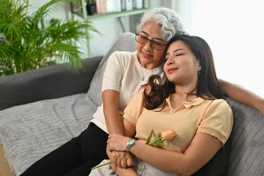 Smiling gray haired mature mother embracing grownup daughter, relaxing on sofa at home.