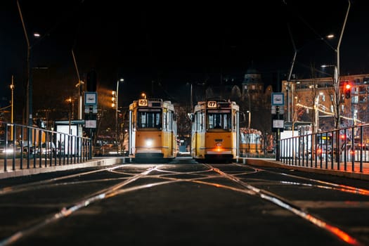 Resting trams locate on rails at night time. Tramways halt on empty thoroughfare illuminated by glow of streetlight waiting for passengers in Luxemburg