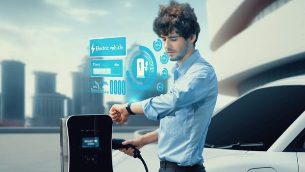 Businessman check EV car battery status on smartwatch hologram while recharge from charging station with modern building background. Futuristic lifestyle of clean energy for EV tech adaptation. Peruse