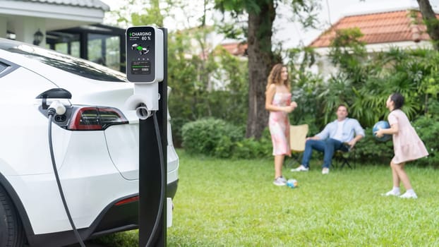 Focus electric vehicle recharge from home charging station on blur background of happy and playful family playing together. EV car using alternative and sustainable energy for better future.Synchronos