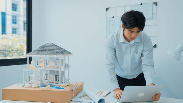 Closeup image of professional architect engineer using laptop analysis data on meeting table with house model and map scatter around at room with blueprint hanged behind. Focus on hand. Immaculate.