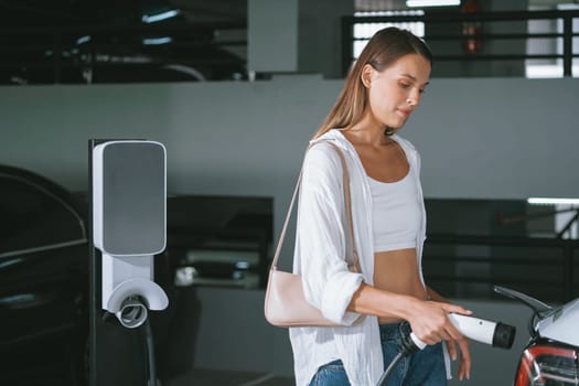 Young woman travel with EV electric car to shopping center parking lot charging in downtown city showing urban sustainability lifestyle by green clean rechargeable energy of electric vehicle innards