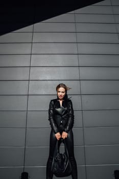 Beautiful fashionable woman in black leather clothes standing against dark wall walk