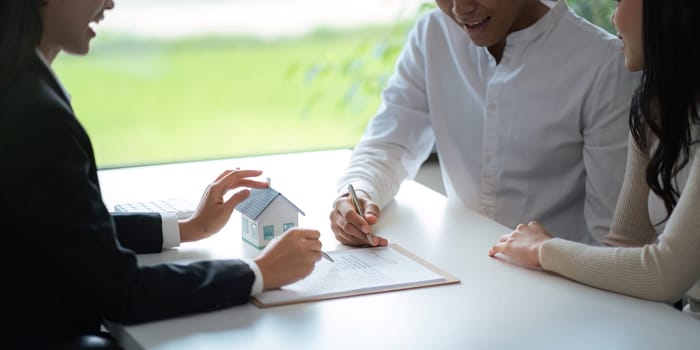 Young Asian couple making contract with house sale agency. man and his wife sitting signing the contract next to him looking the contract document with smile. real estate agreement successful concept.