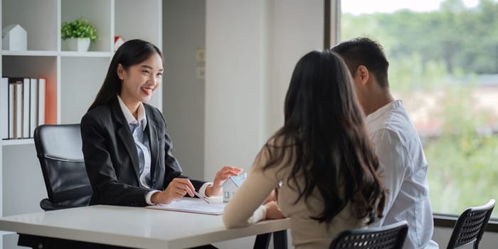 Young Asian couple making contract with house sale agency. man and his wife sitting signing the contract next to him looking the contract document with smile. real estate agreement successful concept.