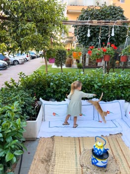 Little girl trying to pet a ginger cat jumping on a mat in the garden. High quality photo