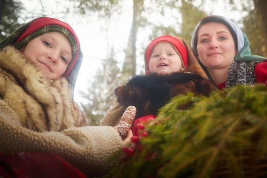 Family with mother, teenage girl, and little daughter dressed in stylized medieval peasant clothing in winter forest. Woman and her daughters pose for fairytale photoshoot in nature on a cold day