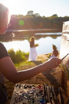 Adult female artist painting picture near water of river or lake in nature and girls in white sundress and flower wreath. Artist and models posing in holiday of Ivan Kupala in nature at sunset