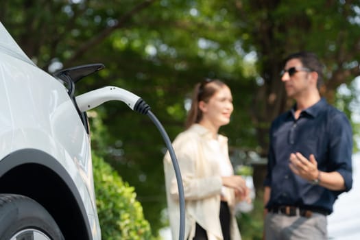 Young couple recharge electric car battery from charging station in green city park in springtime. Rechargeable EV car for sustainable environmental friendly urban travel lifestyle. Panorama Expedient