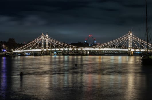 London, UK - Nov 20, 2023 - Nocturnal view of floodlit Albert Bridge from Chelsea Bridge with the reflection of the light in very calm water of River Thames. Lit up at Night Illuminated Albert bridge in west London at night. Space for text, Selective Focus.