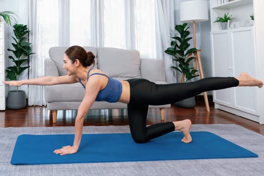 Asian woman in sportswear doing yoga exercise on fitness mat as her home workout training routine. Healthy body care and calm meditation in yoga lifestyle with comfortable and relaxation. Vigorous