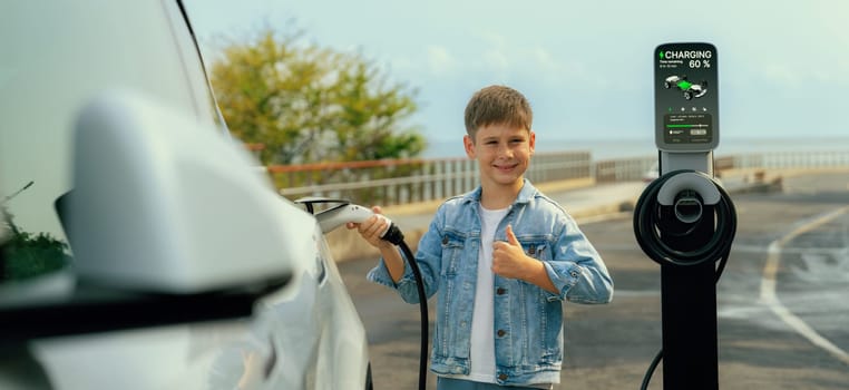 Little boy recharging eco-friendly electric car from EV charging station. EV car road trip travel by the seashore using clean renewable and sustainable energy. Panorama Perpetual