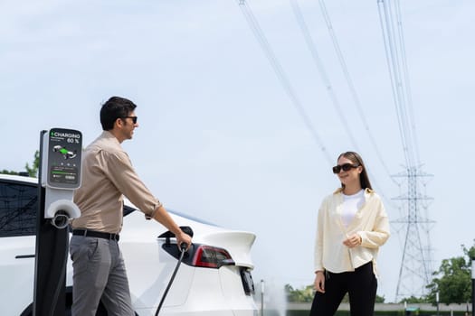 Young couple recharge EV car battery at charging station connected to power grid tower electrical industrial facility as electrical industry for eco friendly vehicle utilization. Expedient