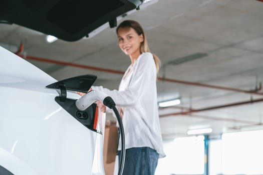 Young woman travel with EV electric car to shopping center parking lot charging in downtown city showing urban sustainability lifestyle by green clean rechargeable energy of electric vehicle innards