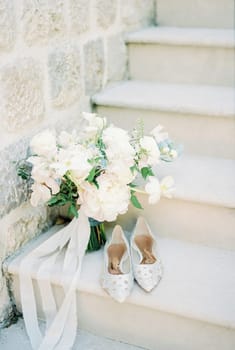 Bride shoes stand on the stone steps of an ancient house near a bouquet of flowers. High quality photo