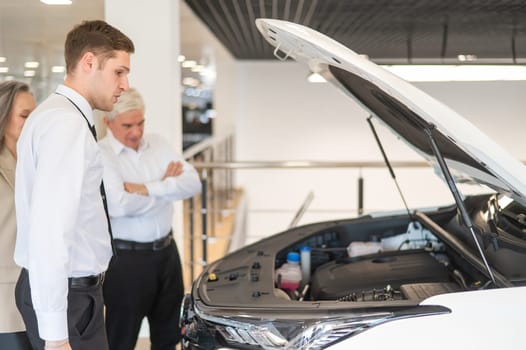 A salesman demonstrates a car with an open hood to an elderly Caucasian couple in a car dealership