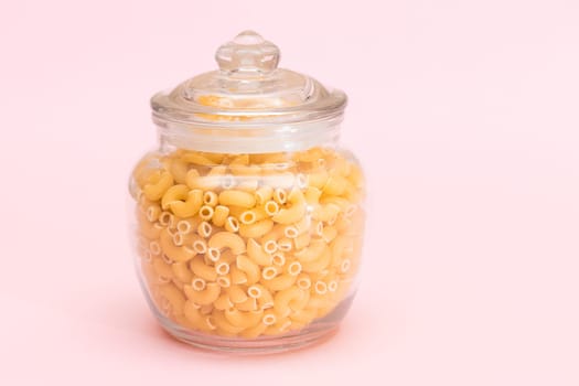 Uncooked Chifferi Rigati Pasta in Glass Jar on Pink Background. Fat and Unhealthy Food. Scattered Classic Dry Macaroni. Italian Culture and Cuisine. Raw Pasta