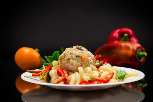 cooked pasta with meatballs, peppers and herbs with spices in a plate, isolated on a black background