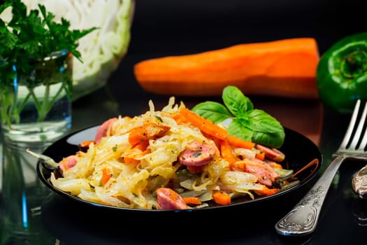 cooked fried cabbage with vegetables and sausages, in a plate isolated on a black background