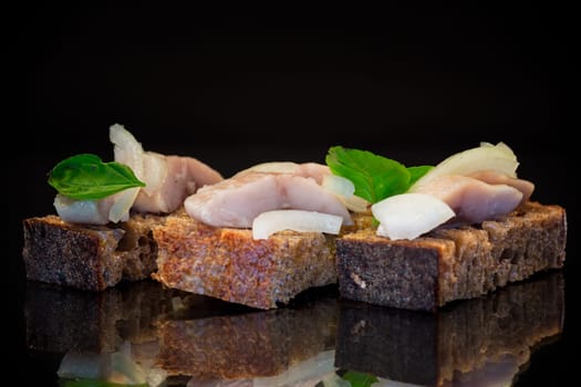canape of pieces of salted herring with onions on a fresh dark piece of bread, isolated on a black background.