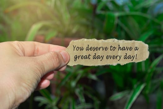 Hand holding torn paper with text on blurred green background - You deserve to have a great day every day.