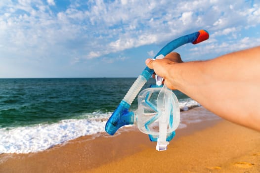 Blue diving mask and snorkel on a sea background. Snorkeling equipment in the hand of a man on a beach holiday while swimming in the ocean water, a break on the shore. Summer or winter direction.