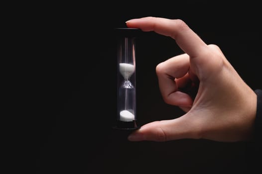 Close-up of a woman holding an hourglass in the dark. Fingers hold on both sides a plastic sand clock with white sand on a black background.