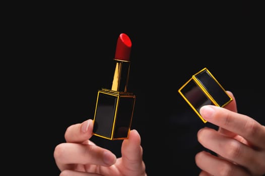 A woman's hand holding a red lipstick in a gold-black frame in one hand, in the other hand a lipstick cap, close-up on a black background.