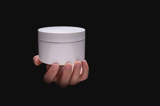 White cosmetic packaging mockup. A woman's hand holds a round jar of body cream on a black background. Beauty industry concept. Close-up, copy space, banner.