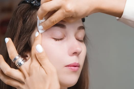 A girl washes paint off her eyebrow. A female cosmetologist wipes makeup off a client's face with a cotton pad. Cosmetologist, makeup concept, beauty salon plan