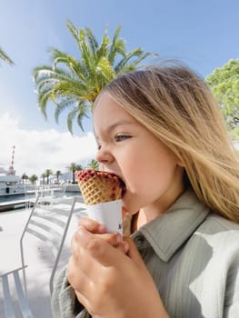 Little girl bites ice cream in a waffle cone while sitting on a chair in a street cafe. High quality photo
