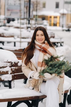 A girl with long hair in winter sits on a bench outside with a bouquet of fresh fir branches.