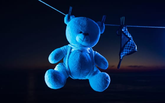 teddy bear toy hangs on a clothesline on clothespins in neon light. tied with shibari ropes