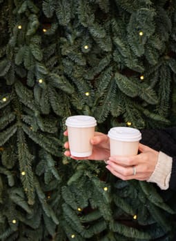 Two hands holding paper cups of coffee in front of a Christmas tree